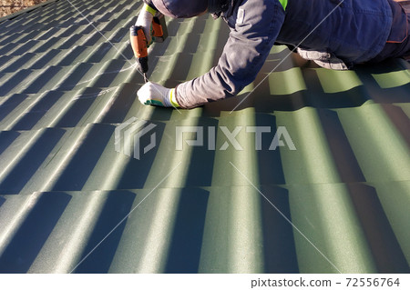 The professional worker works on installation of a roof of a roof by sheets of a metal tile and drills a screw with a drill 72556764