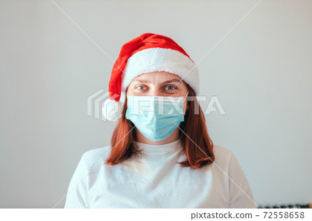 Close up of girl in santa hat and protective medical white mask on grey wall background. New Year in quarantine during the coronavirus pandemic 72558658