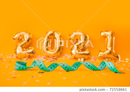 Green measuring tape or tape measure on orange background from 2021 foil balloons with confetti. Preparation for celebrating new year or christmas, weight loss, fitness concept Green measuring tape or tape measure on orange background from 2021 foil balloons with confetti. Preparation for celebrating new year or christmas, weight loss, fitness concept 72558661