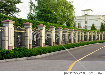 a stone fence with a rustik and an iron forged insert with a pattern and peaks near asphalt road along the fence and hedges of evergreen bushes. a stone fence with a rustik and an iron forged insert with a pattern and peaks near asphalt road along the fence and hedges of evergreen bushes. 72559367