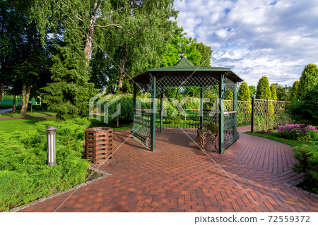 an iron gazebo with a bench and paving stone tiles in the garden with a lawn and evergreen thuja near the canopy of the urn and in the bushes an iron lantern on a sunny day with a cloudy sky, nobody. an iron gazebo with a bench and paving stone tiles in the garden with a lawn and evergreen thuja near the canopy of the urn and in the bushes an iron lantern on a sunny day with a cloudy sky, nobody. 72559372
