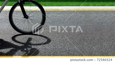 A bicycle wheel rides along an asphalt road with yellow markings and green grass on the sidelines on a sunny summer day with copy space, closeup 16:9. A bicycle wheel rides along an asphalt road with yellow markings and green grass on the sidelines on a sunny summer day with copy space, closeup 16:9. 72560324