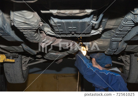 A mechanic with a lamp examines the bottom of the car 72561025