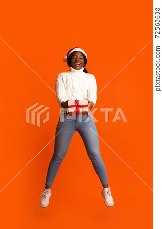 Cheerful black woman in santa hat jumping with christmas present in hands 72563638
