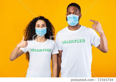 Volunteers Pointing Fingers At Masks Gesturing Thumbs-Up Over Yellow Background Volunteers Pointing Fingers At Masks Gesturing Thumbs-Up Over Yellow Background 72563784