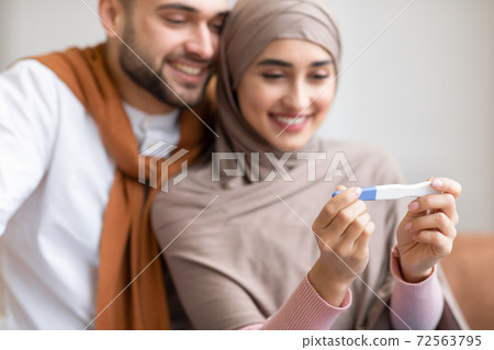 Joyful Muslim Couple Holding Pregnancy Test Sitting Together Indoors 72563795
