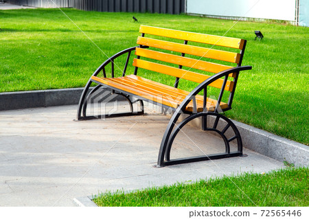 bench with iron black legs and a brown colored wooden seat on a stone sidewalk in the background behind a curb green grass lawn, empty nobody. 72565446