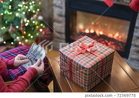 Woman putting american dollars in red christmas sock sitting near christmas tree and fireplace and packing gift box. Woman putting american dollars in red christmas sock sitting near christmas tree and fireplace and packing gift box. 72569778