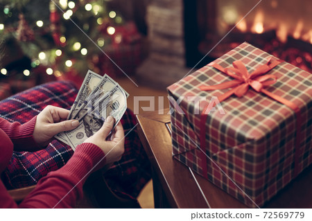Woman putting american dollars in red christmas sock sitting near christmas tree and fireplace and packing gift box. Woman putting american dollars in red christmas sock sitting near christmas tree and fireplace and packing gift box. 72569779
