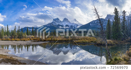 Snow capped The Three Sisters trio peaks mountain with blue sky and white clouds reflect on water surface in autumn. Beautiful natural scenery landscape at Canmore, Canadian Rockies, Alberta, Canada. 72569970