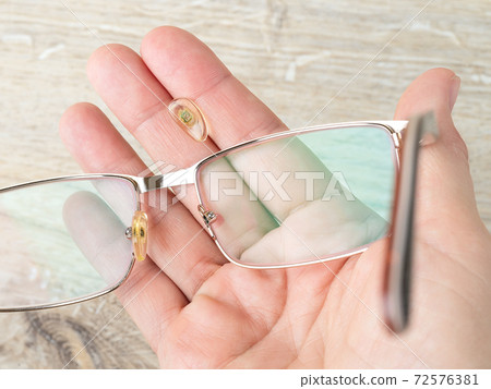Eyeglasses and nose pad broke off them on a hand palm against brown desk. Eyeglasses and nose pad broke off them on a hand palm against brown desk. 72576381