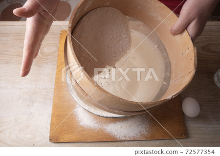 Large bowl, cutting board, sieve for sifting flour on the table. Female hands sifting flour in the home kitchen. 72577354