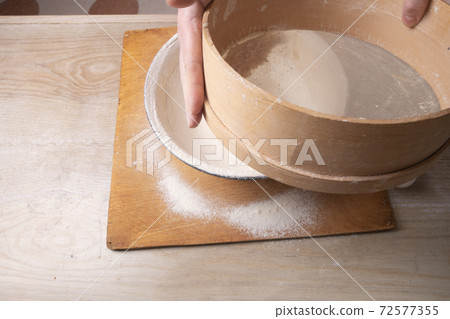 Large bowl, cutting board, sieve for sifting flour on the table. Female hands sifting flour in the home kitchen. Large bowl, cutting board, sieve for sifting flour on the table. Female hands sifting flour in the home kitchen. 72577355