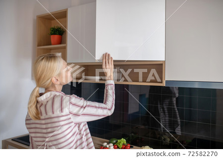 Blonde woman opening the cupboard in the kitchen 72582270
