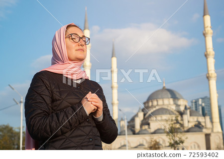 Young woman in a pink scarf and glasses whispers something while holding her hands on the background of the mosque 72593402