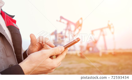 Close-up of a male hand in uniform with a phone on the background of pumping oil Close-up of a male hand in uniform with a phone on the background of pumping oil 72593434