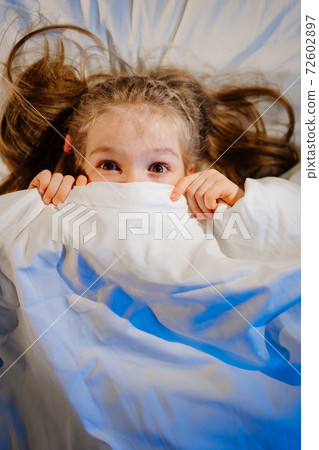 top View. Little girl lies in bed, frightened and covers face with blanket top View. Little girl lies in bed, frightened and covers face with blanket 72602897