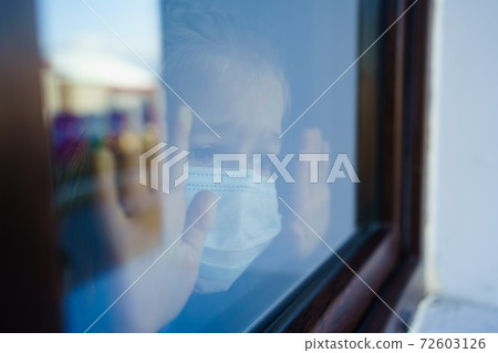 through glass. little girl in protective mask, look out of window outside.  72603126