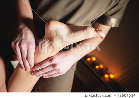 A man masseur does a massage of the thigh and lower leg to a woman client in a dark room of the massage room 72604584