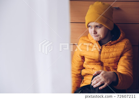 A sad teenager with a yellow jacket and cap sits by the window. A sad teenager with a yellow jacket and cap sits by the window. 72607211
