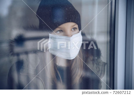 A girl in a black cap with a protective mask stands by the window. A girl in a black cap with a protective mask stands by the window. 72607212