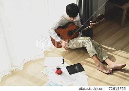 Young man is sitting at home next to the window and playing the acoustic guitar. 72613753