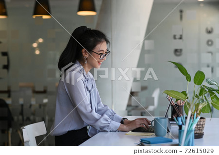 Side view of cheerful young woman sitting in front of her work desk and working with computer tablet. 72615029
