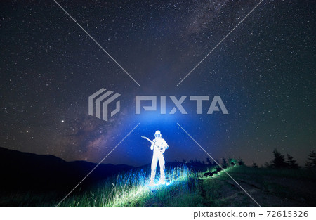 Illuminated spaceman fresh from the space standing in the middle of mountain meadow holding guitar, under Milky Way 72615326