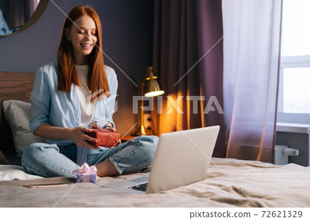 Cheerful young woman holding gift box with present during online communication using laptop. Cheerful young woman holding gift box with present during online communication using laptop. 72621329