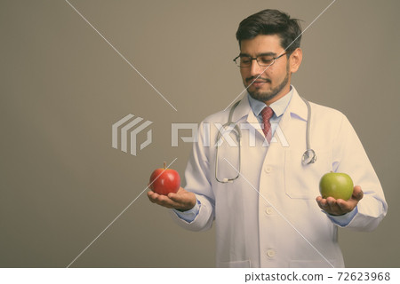 Young handsome bearded Persian man doctor against gray background Young handsome bearded Persian man doctor against gray background 72623968