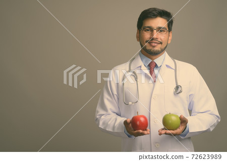 Young handsome bearded Persian man doctor against gray background Young handsome bearded Persian man doctor against gray background 72623989