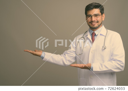 Young handsome bearded Persian man doctor against gray background Young handsome bearded Persian man doctor against gray background 72624033