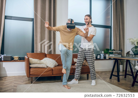 Man in vr glasses walking in the room, his partner supporting him Man in vr glasses walking in the room, his partner supporting him 72632553