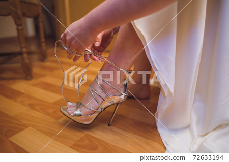 Bride dresses shoes before the wedding ceremony. Charges of the bride. Closeup detail of bride putting on high heeled sandal wedding shoes. Wedding bride shoes. Beautiful legs. Bride dresses shoes before the wedding ceremony. Charges of the bride. Closeup detail of bride putting on high heeled sandal wedding shoes. Wedding bride shoes. Beautiful legs. 72633194