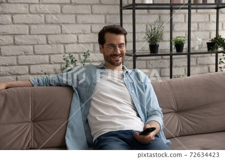 Smiling young guy resting before tv screen holding remote control Smiling young guy resting before tv screen holding remote control 72634423