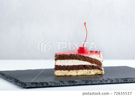 Piece of white and chocolate sponge cake with maraschino cherry on black plate. Side view to piece of cake Piece of white and chocolate sponge cake with maraschino cherry on black plate. Side view to piece of cake 72635537