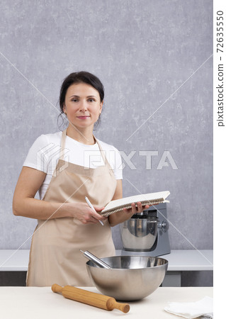 Portrait of woman chef in kitchen with cookbook in her hands. Bowl and rolling pin on table. Vertical frame 72635550