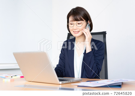 一個戴著中年眼鏡的女人在辦公室會議室在智能手機上聊天時在計算機上工作 一個戴著中年眼鏡的女人在辦公室會議室在智能手機上聊天時在計算機上工作 72645820