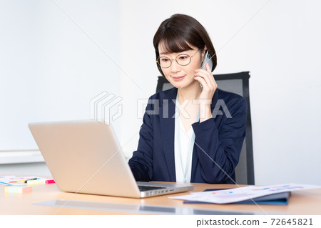 一個戴著中年眼鏡的女人在辦公室會議室在智能手機上聊天時在計算機上工作 72645821