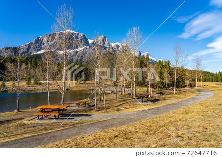 Quarry Lake park lakeshore in late autumn season sunny day morning. Snow capped Mount Rundle mountains in with blue sky in the background. Canmore, Alberta, Canada. Quarry Lake park lakeshore in late autumn season sunny day morning. Snow capped Mount Rundle mountains in with blue sky in the background. Canmore, Alberta, Canada. 72647716