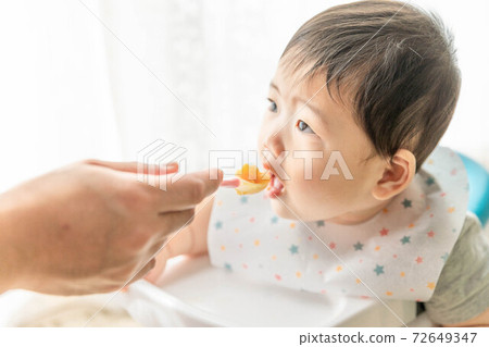 Baby sitting on a baby chair by the window and eating baby food 72649347
