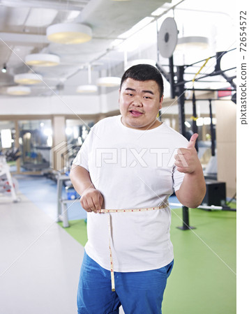 asian overweight man measuring waist and happy with progress in gym asian overweight man measuring waist and happy with progress in gym 72654572
