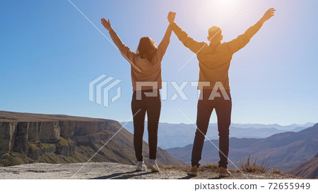 Couple walks and raises joined hands on cliff under blue sky Couple walks and raises joined hands on cliff under blue sky 72655949