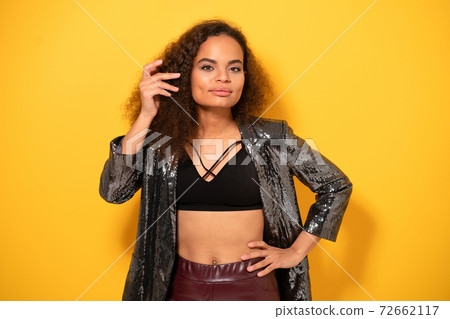 Posh beauty ethnic model girl with beautiful hairstyle smile looking at camera wearing shiny black jacket. Happy young girl fixing hair standing on yellow background. Studio shot Posh beauty ethnic model girl with beautiful hairstyle smile looking at camera wearing shiny black jacket. Happy young girl fixing hair standing on yellow background. Studio shot 72662117