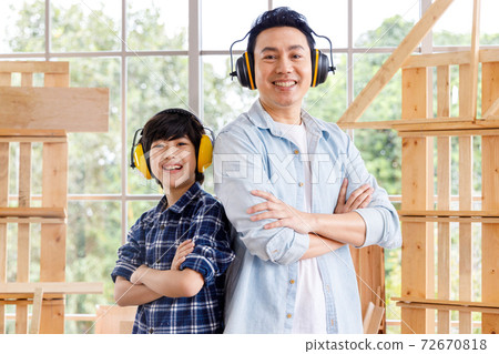 Happy Asian father and son wearing ear muffs and standing in carpentry workshop. Happy Asian father and son wearing ear muffs and standing in carpentry workshop. 72670818
