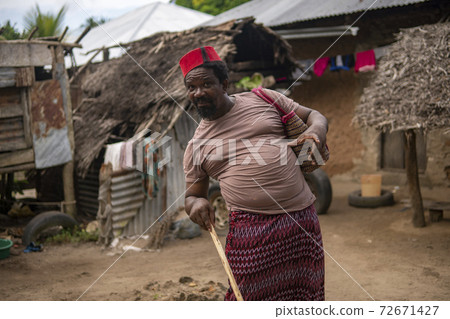 An African Older Man in Red Muslim Taqiyyah Fez Hat posing with a stick for lame people on Yard Near the Basic Hut with Thatched roof in Small Remote Village in Tanzania, Pemba island, Zanzibar 72671427