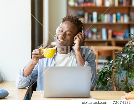 Handsome black guy with headphones listening to music with closed eyes and drinking coffee at cozy cafe 72674182
