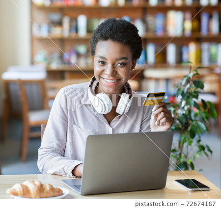 Portrait of happy young black woman with headphones and credit card using laptop for online shopping at cafe Portrait of happy young black woman with headphones and credit card using laptop for online shopping at cafe 72674187