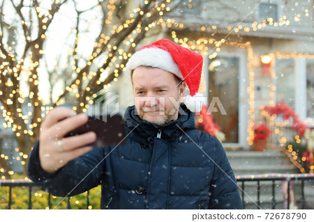 Mature man wearing Santa hat making selfie near his decorated home. Mature man wearing Santa hat making selfie near his decorated home. 72678790