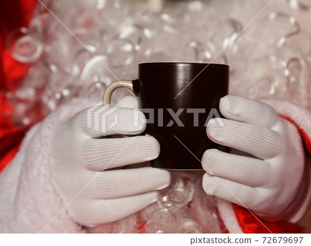 Santa Claus holding hands in a dark brown mug of tea in front of a white beard Santa Claus holding hands in a dark brown mug of tea in front of a white beard 72679697